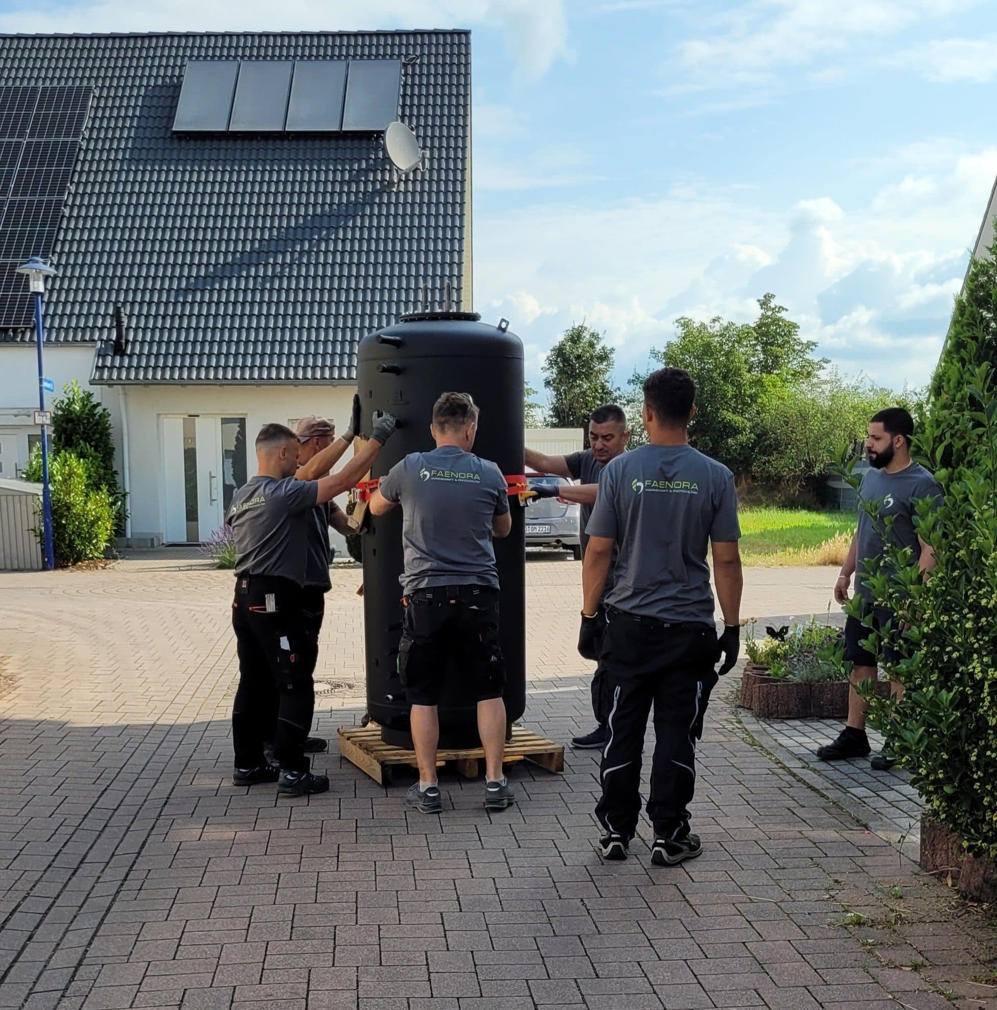 Mehrere Monteure von FAENORA tragen gemeinsam einen großen Pufferspeicher auf einer Palette über die Einfahrt eines Einfamilienhauses