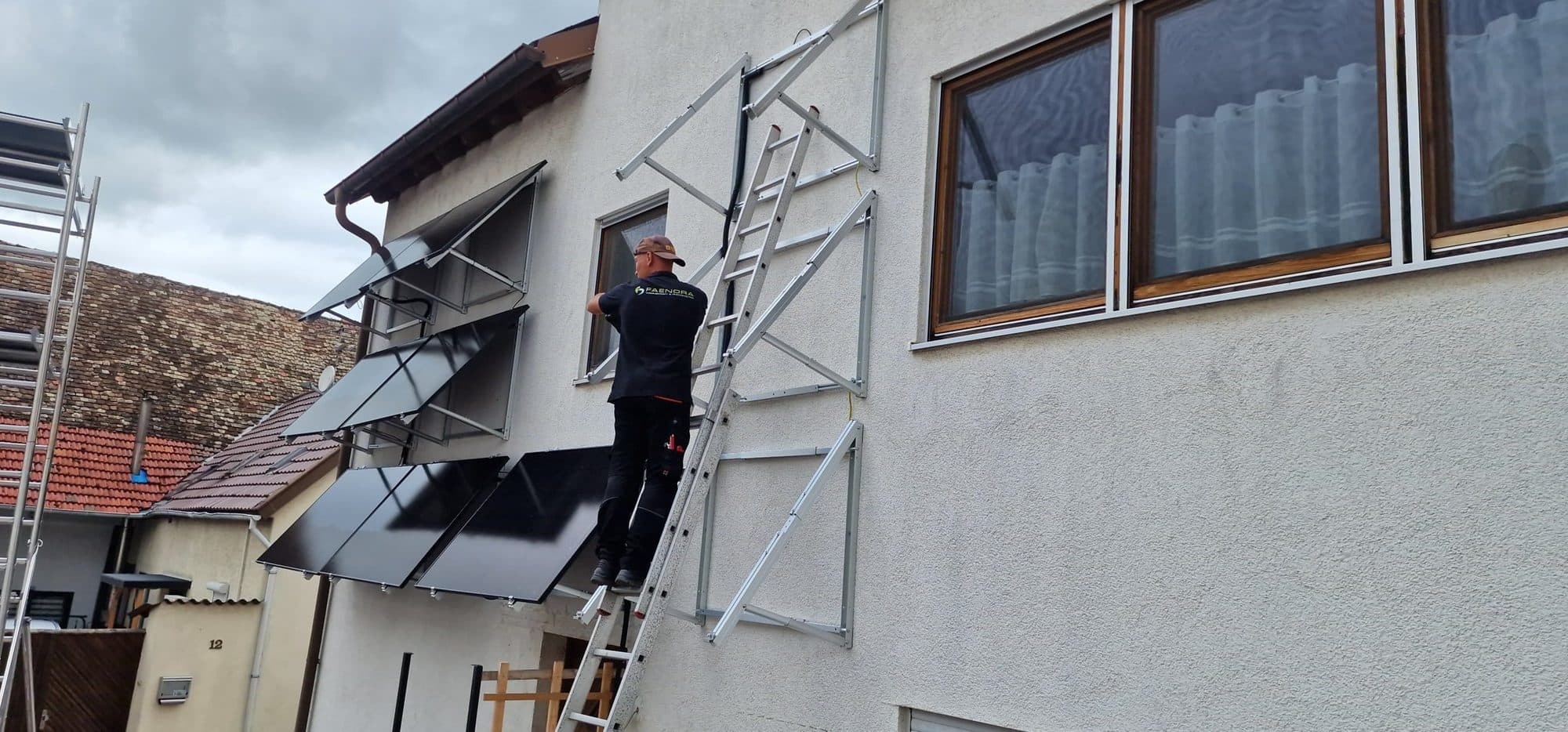 Monteur auf einer Leiter montiert Kollektoren an einer Hausfassade, daneben ein Gerüst