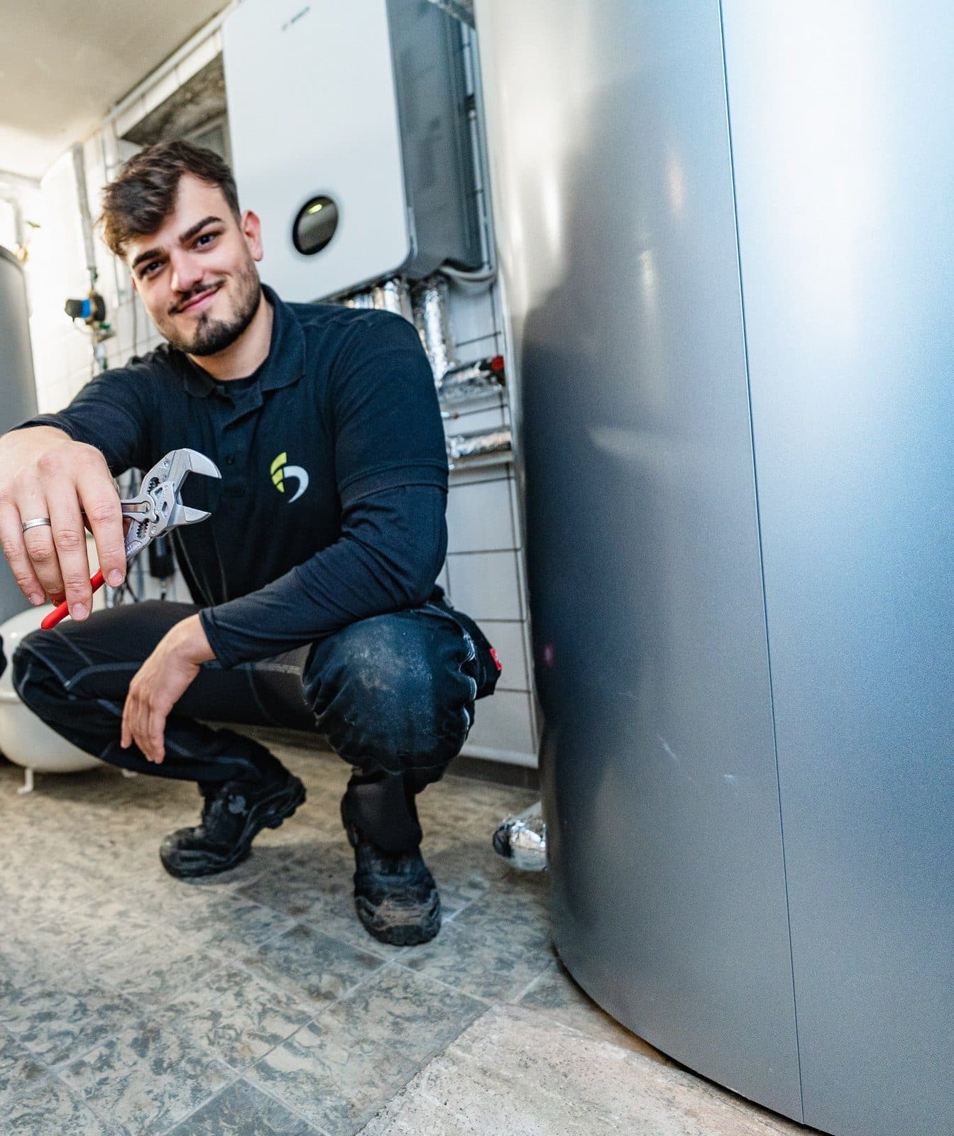Monteur von FAENORA hockt mit einer Zange in der Hand vor einem Warmwasserspeicher