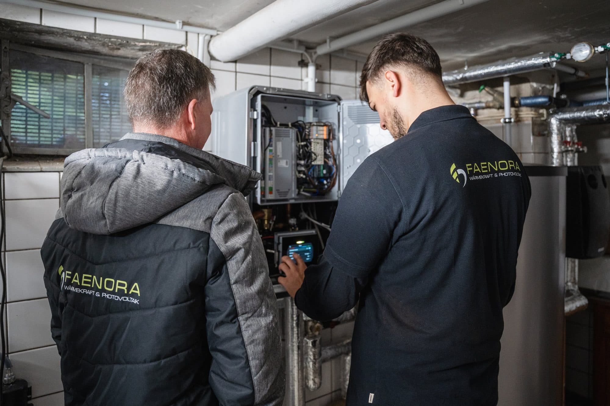 Zwei Monteure von FAENORA in Firmenjacken nehmen im Technikraum eine Wärmepumpen-Inneneinheit in Betrieb